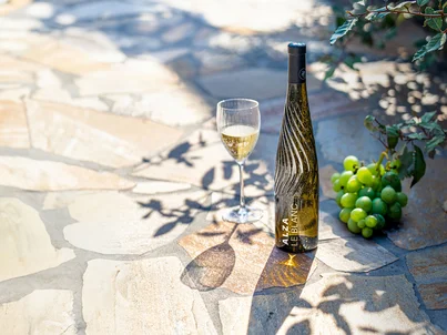 Eine Flasche ALZA Le Vin und ein Weinglas daneben. Das originelle Flaschendesign glitzert im Sonnenlicht.