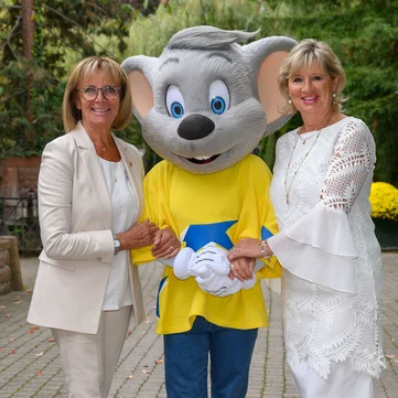 Marianne und Mauritia Mack mit Ed Euromaus vor grüner Kulisse mit Bäumen und Blumen.