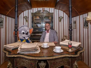 Roland Mack sitzt gemeinsam mit einem großen Plüsch Ed an einem Schreibtisch in einem historischen Zirkuswagen.