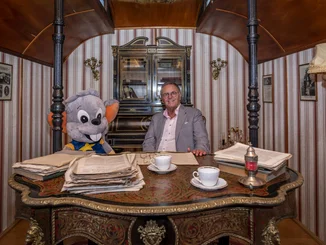 Roland Mack sitzt gemeinsam mit einem großen Plüsch Ed an einem Schreibtisch in einem historischen Zirkuswagen.