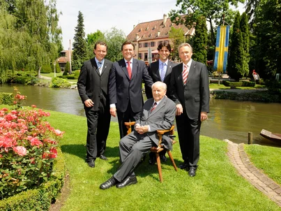 Franz Mack sitzt auf einer Wiese im Europa-Park. Roland, Michael, Thomas und Jürgen Mack stehen hinter ihm, im Hintergrund befindet sich das Schloss Balthasar.