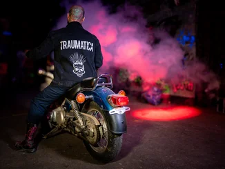 Mann trägt Traumatica Jeansjacke mit Aufschrift, sitzt auf Motorrad.